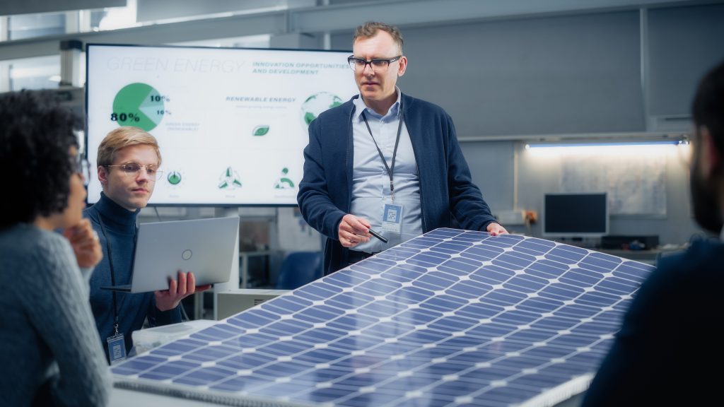 Descrição da imagem oculta: A imagem mostra um ambiente moderno de laboratório ou sala de pesquisa com iluminação artificial. No centro, um homem de meia-idade, branco, com óculos, cabelos curtos e roupa social, veste um crachá e segura uma caneta enquanto apresenta um grande painel solar apoiado sobre uma mesa. Ao redor da mesa, há três outras pessoas. À esquerda, um jovem de cabelos loiros e óculos segura um laptop aberto; no canto esquerdo, uma mulher com cabelo cacheado observa a apresentação com a mão no queixo e, no canto direito da imagem, outra pessoa, parcialmente visível, também participa da reunião. No fundo, há um grande monitor exibindo gráficos e textos sobre energia renovável, com palavras como “Green Energy” e imagens de turbinas eólicas e painéis solares. Fim da descrição