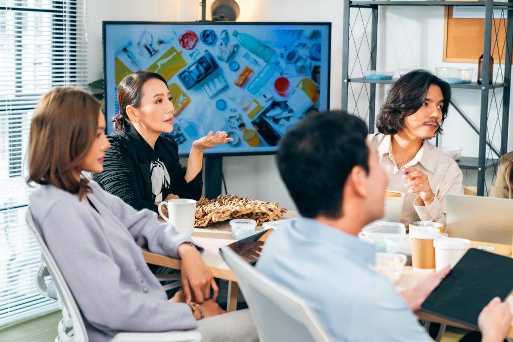 descrição da imagem: A imagem mostra um grupo de quatro pessoas reunidas em uma sala de reunião moderna. No centro da cena, uma mulher de pele clara e cabelos presos, vestindo um blazer preto e uma blusa com laço, gesticula com a mão enquanto fala. À sua frente, uma mulher de cabelos castanhos soltos veste um casaco lilás e está sentada, ouvindo a conversa. Um homem de cabelos escuros e curtos, vestindo uma camisa azul-clara, segura um tablet e olha para a mulher que está falando. Outro homem, com cabelos médios e ondulados, veste uma camisa bege e segura uma caneta, parecendo atento à discussão. A mesa onde o grupo está reunido contém xícaras, potes plásticos com comida e um objeto trançado feito de material natural. Ao fundo, há uma tela grande exibindo uma imagem de objetos coloridos dispostos sobre uma superfície branca, incluindo embalagens, garrafas e utensílios diversos. A sala possui uma estante metálica com caixas e objetos organizados, além de uma janela com persianas, que permite a entrada de luz natural. Fim da descrição