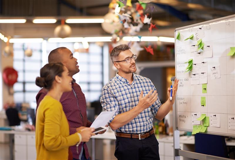 Descrição da imagem: A imagem mostra três pessoas em um ambiente de escritório moderno, discutindo em frente a um quadro branco cheio de anotações e post-its verdes. Um homem branco, de óculos, barba curta e cabelos grisalhos, vestindo uma camisa xadrez azul e branca, segura um marcador e gesticula enquanto fala. Ao seu lado, um homem negro, com a cabeça raspada e vestindo uma camisa vinho com pequenos padrões, observa o quadro com atenção. À frente deles, uma mulher negra de cabelos presos em um coque, vestindo um cardigã amarelo, segura um caderno e uma caneta enquanto acompanha a explicação. O escritório tem um ambiente aberto, com mesas ao fundo, luminárias pendentes e janelas grandes que permitem a entrada de luz natural. No teto, há enfeites coloridos suspensos, incluindo origamis de pássaros. Fim da descrição