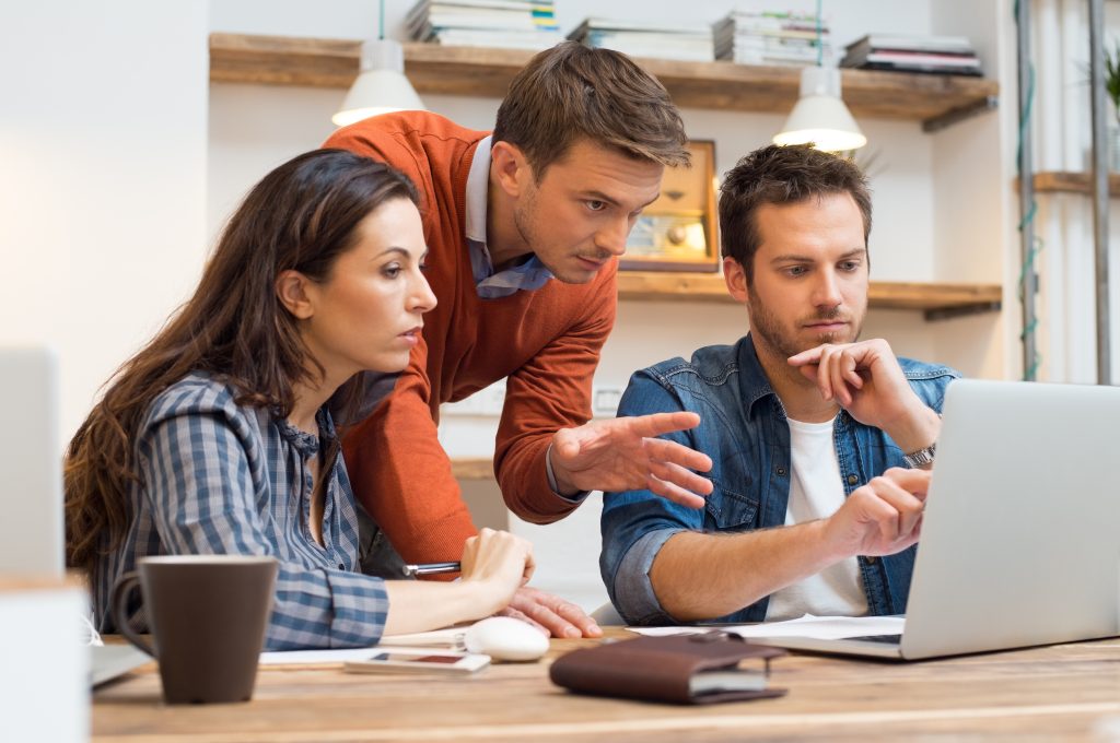 Descrição da imagem: a imagem mostra três pessoas reunidas em um ambiente de escritório, concentradas em uma tela de laptop. Um homem branco, de cabelos curtos e barba, vestindo uma jaqueta jeans sobre uma camiseta branca, está sentado e digitando no computador. Ao seu lado, uma mulher branca de cabelos longos e escuros, vestindo uma camisa xadrez azul e branca, observa a tela com atenção. Atrás deles, um homem branco de cabelos curtos, vestindo um suéter laranja sobre uma camisa azul, inclina-se para a frente, gesticulando com as mãos enquanto parece explicar algo. A mesa contém um caderno fechado, uma caneca e um pequeno objeto branco próximo à mão da mulher. O ambiente tem prateleiras de madeira na parede ao fundo, contendo livros e caixas organizadas. Duas luminárias pendentes iluminam o espaço. Fim da descrição.
