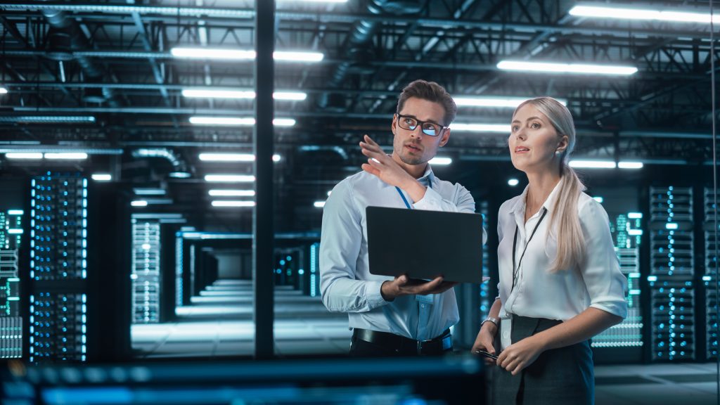 (Descrição da imagem: a imagem mostra dois profissionais, um homem branco e de cabelos curtos e escuros e uma mulher, também branca e de cabelos compridos e loiros, estão em pé dentro de um moderno data center, rodeados por servidores iluminados com luzes azuis. O homem segura um notebook e parece explicar algo com gestos enquanto a mulher escuta atentamente, demonstrando interesse. Ambos usam crachás, sugerindo um ambiente corporativo formal e voltado à tecnologia. A cena transmite uma atmosfera de análise técnica, tomada de decisões e colaboração estratégica, reforçando o papel da inovação e da infraestrutura digital no contexto da governança e transformação organizacional. Fim da descrição).