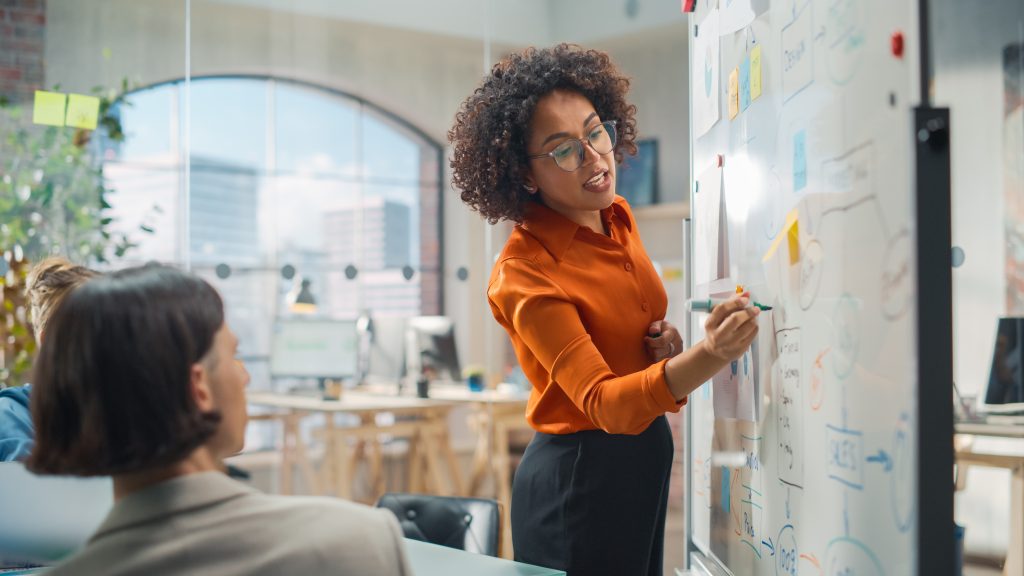 Descrição da imagem: Em um ambiente de escritório iluminado, uma mulher com cabelo cacheado, usando óculos e camisa laranja, está em pé diante de um quadro branco, explicando um conteúdo visual com gráficos, setas e post-its coloridos. Ela segura uma caneta e parece engajada enquanto apresenta suas ideias para um grupo. Na frente dela, parcialmente visíveis, estão colegas sentados, atentos à explicação. Ao fundo, vê-se grandes janelas com vista para prédios urbanos, plantas e estações de trabalho. Fim da descrição.