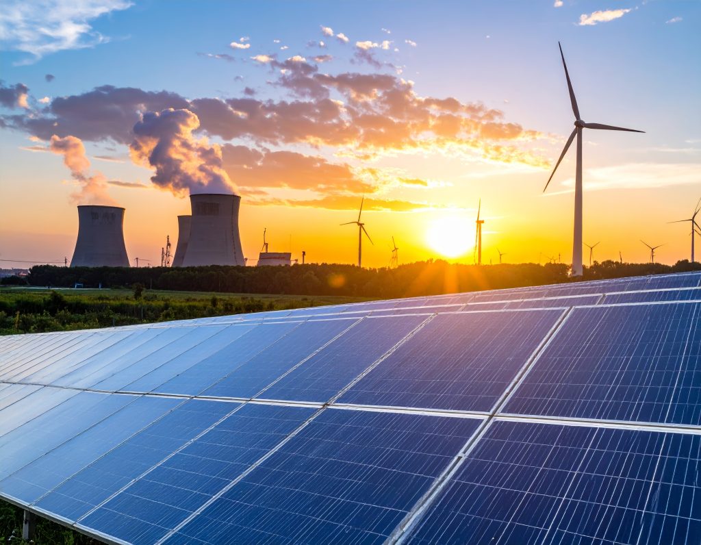 (Descrição da imagem: A imagem exibe uma paisagem ao pôr do sol, com diversas fontes de geração de energia. Em primeiro plano, ocupando a parte inferior da imagem, há uma grande extensão de painéis solares azuis, dispostos em fileiras e inclinados. Ao fundo, contra um céu com nuvens e tons de laranja, amarelo e azul, estão várias estruturas: À esquerda, veem-se três grandes torres de resfriamento de uma usina nuclear, com vapor branco saindo de duas delas. À direita e espalhadas pelo horizonte, há várias turbinas eólicas de cor branca. O sol está se pondo no horizonte, criando um brilho intenso e longas sombras. Entre os painéis solares e as estruturas ao fundo, há uma faixa de vegetação verde. Fim da descrição.)