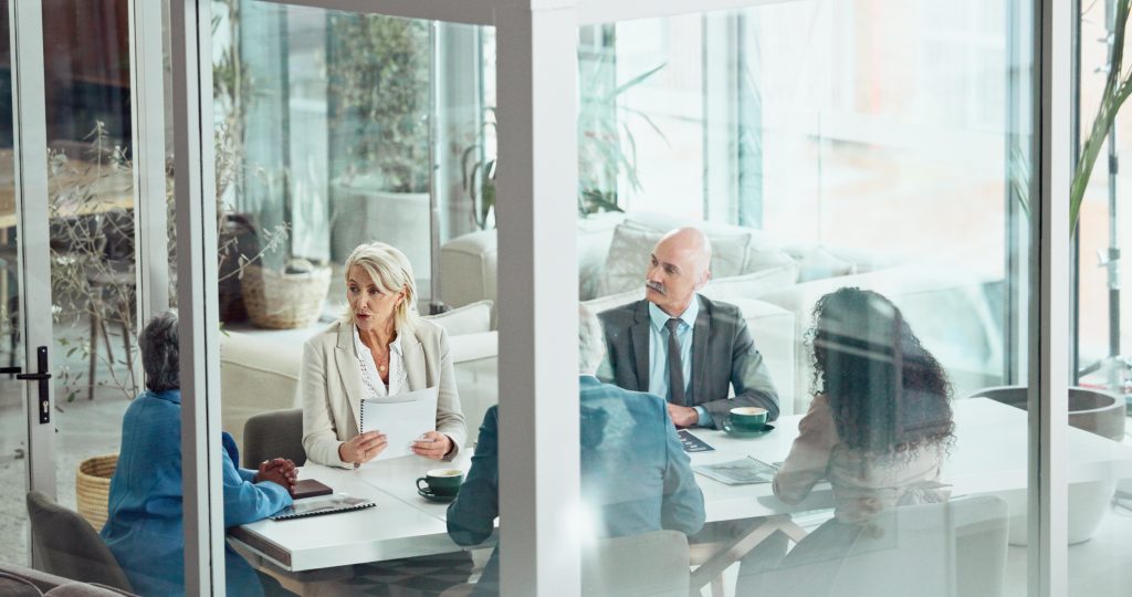 (Descrição da imagem: a imagem mostra um grupo de pessoas em uma reunião de negócios, vistas através de uma divisória de vidro. A cena é dividida ao meio por uma moldura branca da estrutura de vidro. As pessoas estão sentadas ao redor de uma grande mesa branca em um escritório moderno e bem iluminado.
À esquerda, uma mulher com cabelos grisalhos e curtos, vestindo um blazer bege, está sentada à mesa, segurando papéis e falando. Em frente a ela, outra pessoa é vista de costas, vestindo uma roupa azul. À direita, um homem calvo com bigode, vestindo terno e gravata, olha para a mulher que está falando. Ao seu lado, de costas para a câmera, está uma mulher com longos cabelos escuros e cacheados. Outra pessoa, de costas, vestindo um blazer azul, também está sentada à mesa. Sobre a mesa, há xícaras de café, papéis e cadernos. O ambiente ao fundo é claro, com sofás brancos e plantas, visíveis através das paredes de vidro. Fim da descrição).