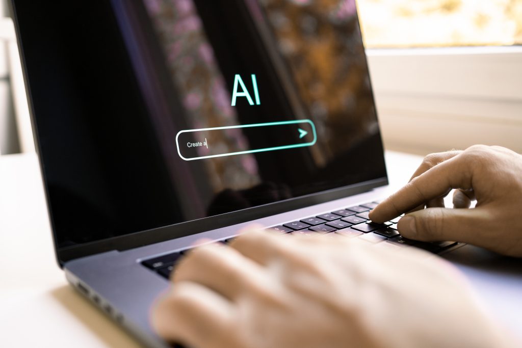 (Descrição da imagem: A imagem mostra as mãos de uma pessoa digitando em um laptop, que está sobre uma mesa clara. O foco principal é a tela do computador. Na tela, que tem um fundo escuro, as letras