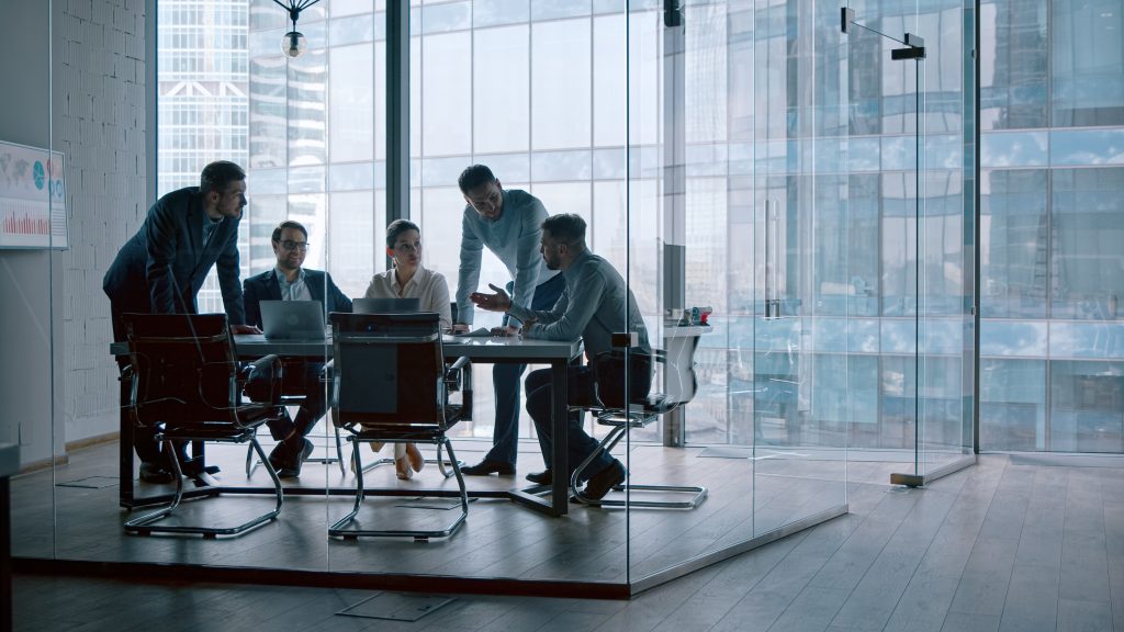 (Descrição da imagem: A imagem mostra um grupo de cinco pessoas em uma reunião dentro de uma sala de conferências com paredes de vidro. As pessoas estão reunidas em torno de uma mesa retangular. Três estão sentadas e duas estão em pé, inclinadas sobre a mesa. Todos estão vestidos com roupas de trabalho, como ternos e camisas. Um homem de terno está em pé na ponta esquerda da mesa, olhando para os papéis. Sentados, há um homem de óculos com um laptop à sua frente e uma mulher ao seu lado.
Outros dois homens estão na outra ponta da mesa; um está em pé, inclinado, e o outro está sentado, gesticulando com a mão enquanto conversa. Na parede à esquerda, há um quadro branco com gráficos coloridos. A sala é cercada por grandes painéis de vidro que vão do chão ao teto, através dos quais se pode ver a fachada de um arranha-céu moderno. O piso da sala é de madeira clara. Fim da descrição).