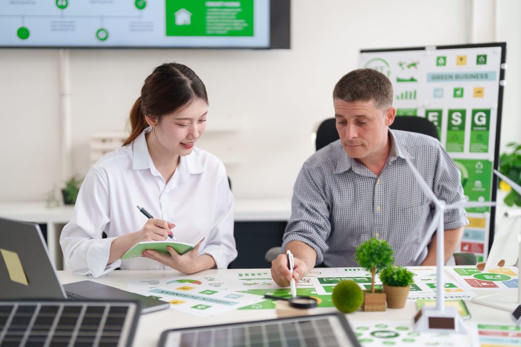 (Descrição da imagem: A imagem mostra um homem e uma mulher sentados lado a lado em uma mesa de escritório, trabalhando em um projeto sobre sustentabilidade. A mulher, à esquerda, é jovem, de ascendência asiática, e veste uma camisa branca. Ela sorri enquanto escreve em um pequeno caderno verde. O homem, à direita, é mais velho, branco, e veste uma camisa xadrez de manga comprida. Ele está inclinado sobre a mesa, analisando alguns papéis. A mesa está coberta com materiais relacionados a energia limpa e negócios sustentáveis. Há papéis com gráficos e ícones verdes, um laptop, um pequeno painel solar, e miniaturas de árvores e uma turbina eólica. Ao fundo, há uma grande tela de TV na parede exibindo um infográfico verde e um quadro branco com gráficos e a sigla