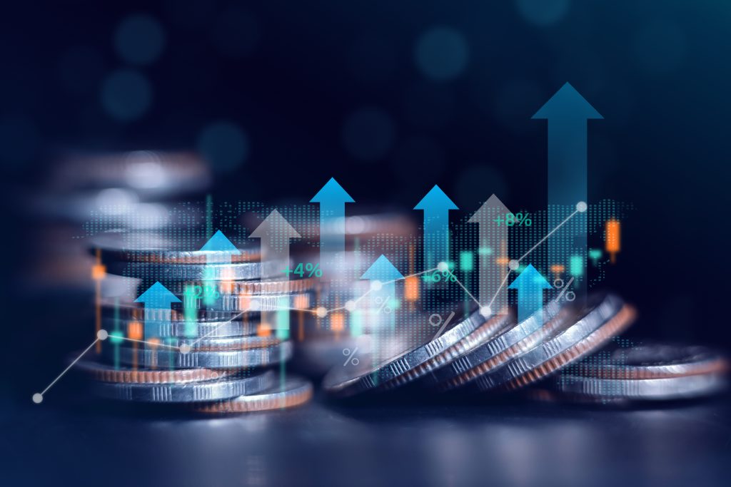 (Descrição da imagem: A imagem apresenta uma composição que combina elementos físicos e digitais para representar crescimento financeiro. Em primeiro plano, há várias pilhas de moedas prateadas de diferentes alturas, dispostas sobre uma superfície escura. Algumas moedas estão perfeitamente empilhadas, enquanto outras aparecem levemente inclinadas ou espalhadas na base das pilhas. Sobrepostas às moedas, há diversas representações gráficas digitais e luminosas, como setas de tamanhos diferentes, em tons de azul e cinza, que apontam verticalmente para cima; Gráfico de Linha, que é uma linha branca fina e ascendente que percorre a imagem da esquerda para a direita, com pequenos pontos circulares marcando os vértices. Temos também Dados Numéricos, que são pequenos textos e símbolos flutuantes, como